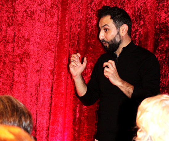 Vor einem dunkelroten Tuch steht eine Person und erzählt gestenreich vor Publikum. Das Publikum sitzt, es sind die Hinterköpfe zu sehen.