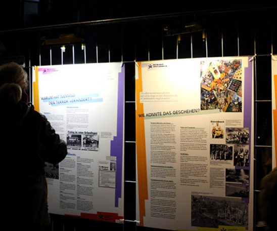 Ein Bild auf einer dunklen Straße: Auf Bauzäunen sind Plakatwände montiert, die von oben mit Strahlern beleuchtet werden. Die Schemen von Personen, die sich die Plakatwände anschauen sind zu sehen.