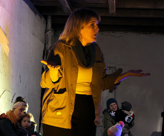 In einem Stall sitzen im Hintergrund Personen mit Kindern auf dem Schoß oder Arm. Im Vordergrund befindet sich eine Person, die ausdrucksstark mit Mimik und Händen gestikuliert und dabei etwas sagt.