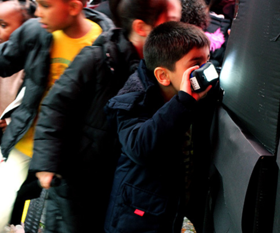 Mehrere kleine Kinder drängeln sich vor schwarzen Boxen. Eins der Kinder hat eine Taschenlampe an und lugt in eine der Boxen hinein.