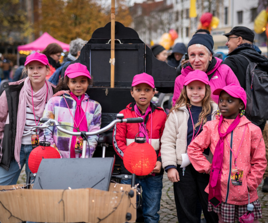 Fünf Kinder in pinker Kleidung und eine Erwachsene schauen in die Kamera. Im Hintergrund ist ein Platz mit Menschen zu sehen und eine schwarze Kiste.