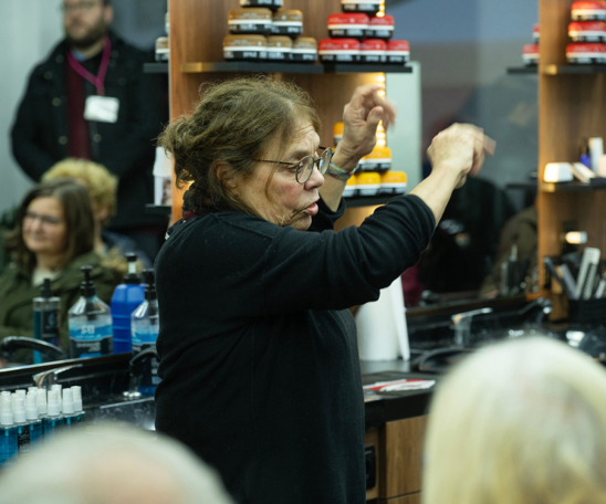 Eine Person gestikuliert ausdrucksstark und spricht dabei. Hinter ihr sind die Spiegel und Regale eines Frisörsalons zu sehen. Im Spiegel sind sitzende und stehende Personen im Raum zu erkennen.