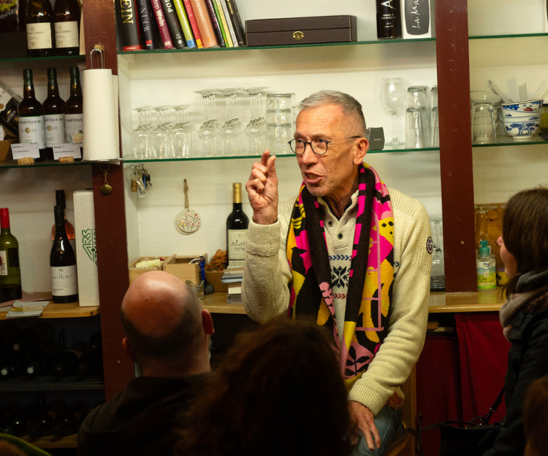 Vor einem Regal mit Gläsern und Weinflaschen und Büchern sitzt eine Person mit Brille und bunten Schal. Vor der Person sind die Köpfe von Zuhörenden zu sehen, die ihn anschauen. Sie hält eine Hand hoch und macht dabei ein Handzeichen und spricht.