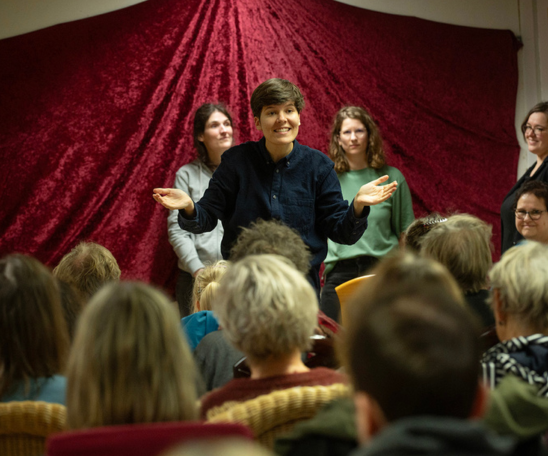 Ein Publikum sitzt uns mit dem Rücken zugewandt vor einer Bühne mit rotem Hintergrund. Vor dem Hintergrund stehen mehrere Personen, eine gestikuliert ausdrucksstark und lächelt das Publikum an.